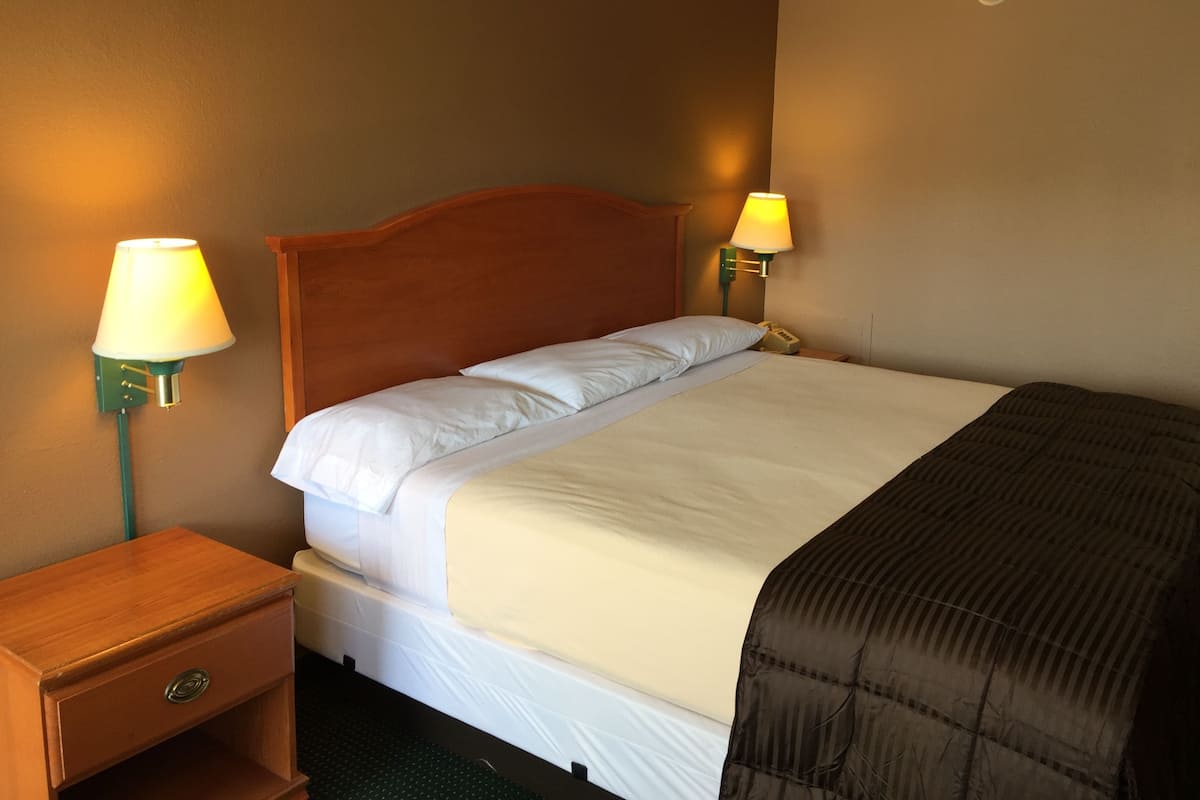 A hotel room with a wooden headboard, a single bed with white bedding, two bedside lamps, a nightstand with a drawer, and a wall-mounted thermostat.