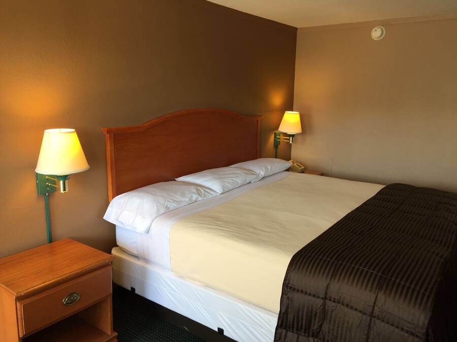 Une chambre d’hôtel avec une tête de lit en bois, un lit simple avec une literie blanche, deux lampes de chevet, une table de chevet avec un tiroir et un thermostat fixé au mur.