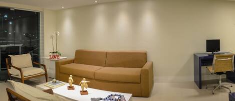 A living room with a beige sofa, a small table with decorative items, a chair, a side table with a plant, a desk with a computer, and a decorative metal sculpture.
