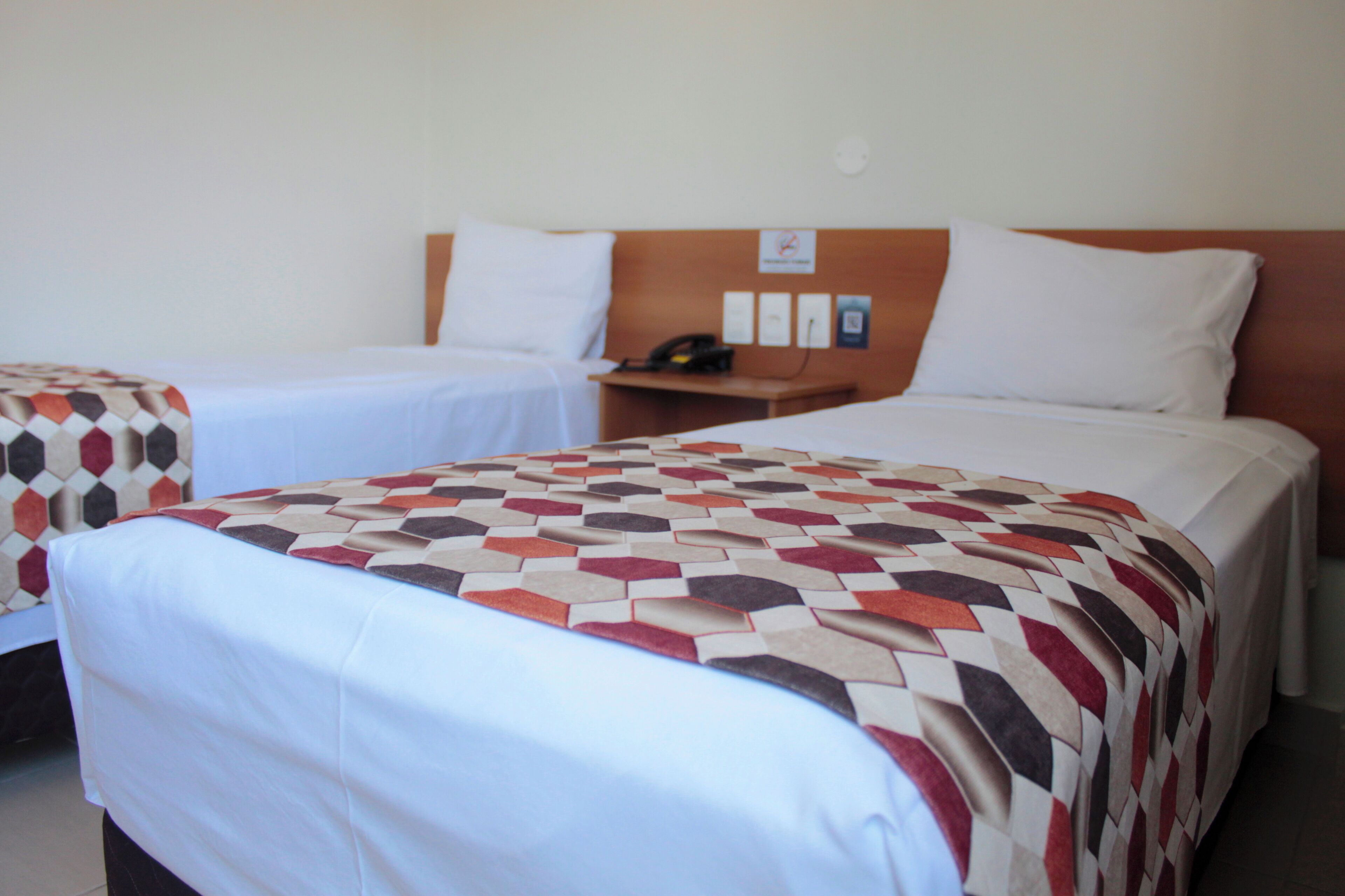 A hotel room with two single beds, a wooden headboard, a bedside table with a phone, and a wall-mounted light switch.