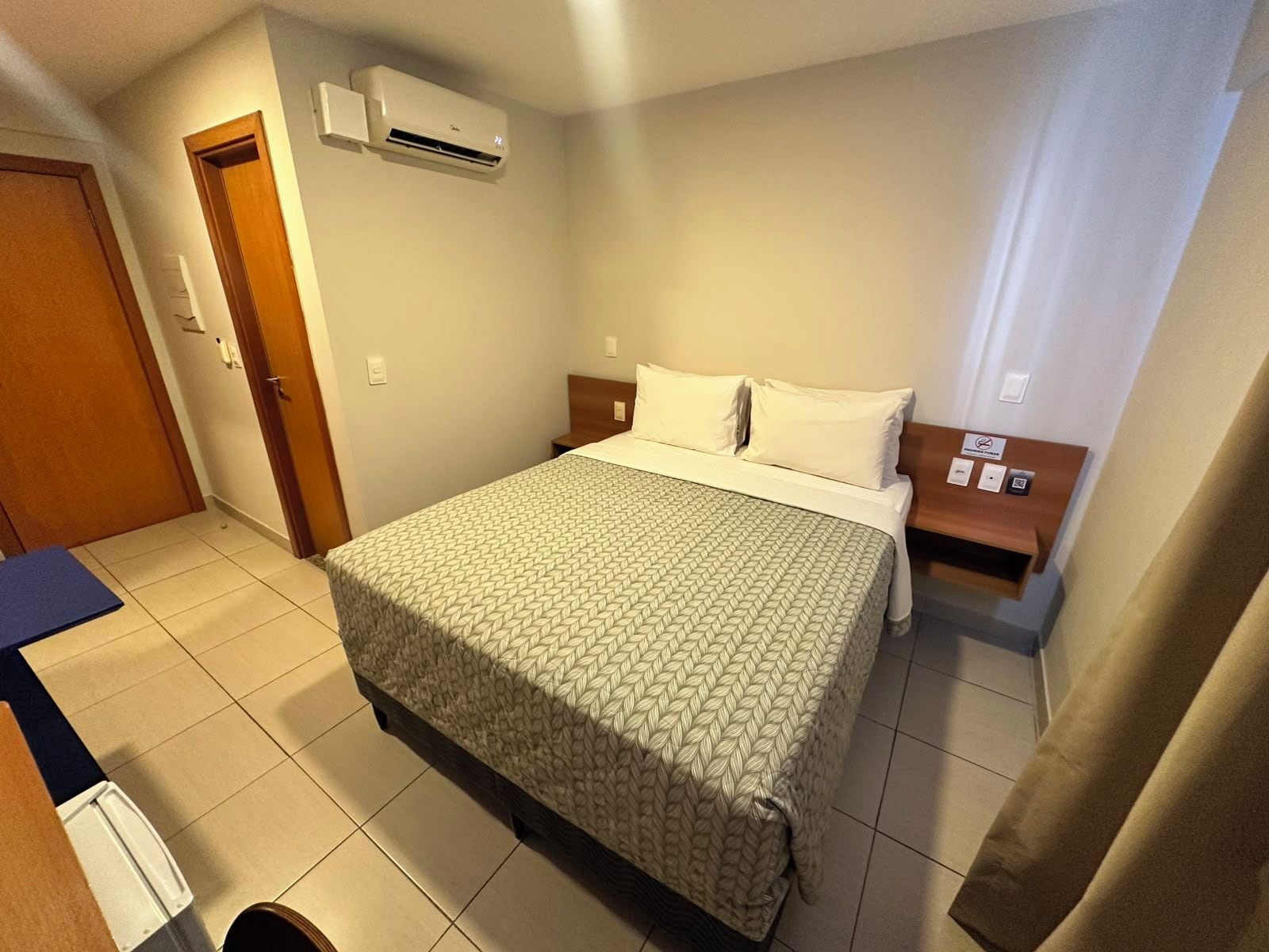 A hotel room with a bed, a nightstand, an air conditioning unit, a door, and a bathroom.