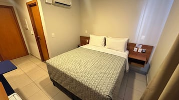 Una habitación de hotel con cama, mesita de noche, equipo de aire acondicionado, puerta y baño.