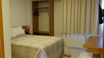 Quarto de hotel com cama, guarda-roupa de madeira, mesinha e frigorífico branco.