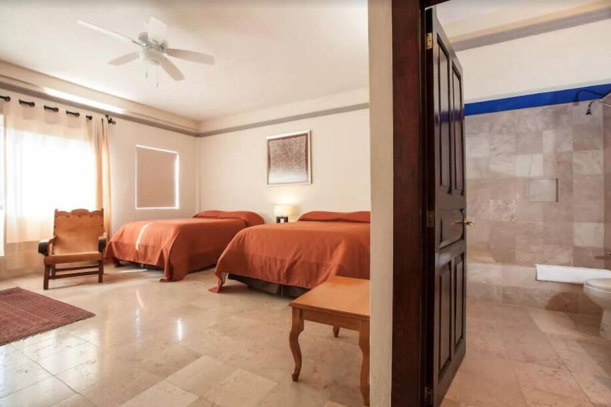 Une chambre d’hôtel avec deux lits, une chaise, une table, un ventilateur de plafond et une salle de bain avec douche et toilettes.