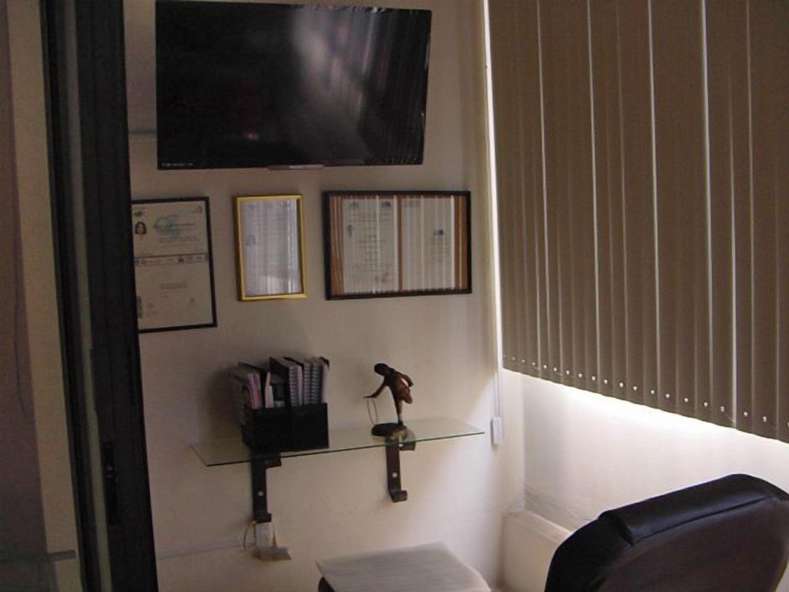 Una habitación con un televisor de pared, diplomas enmarcados, un estante pequeño con libros y un objeto decorativo, y una ventana con persianas.