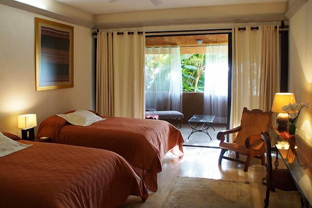 Une chambre d’hôtel avec deux lits, un ventilateur de plafond, une table avec une lampe et une vue sur un balcon avec des rideaux.