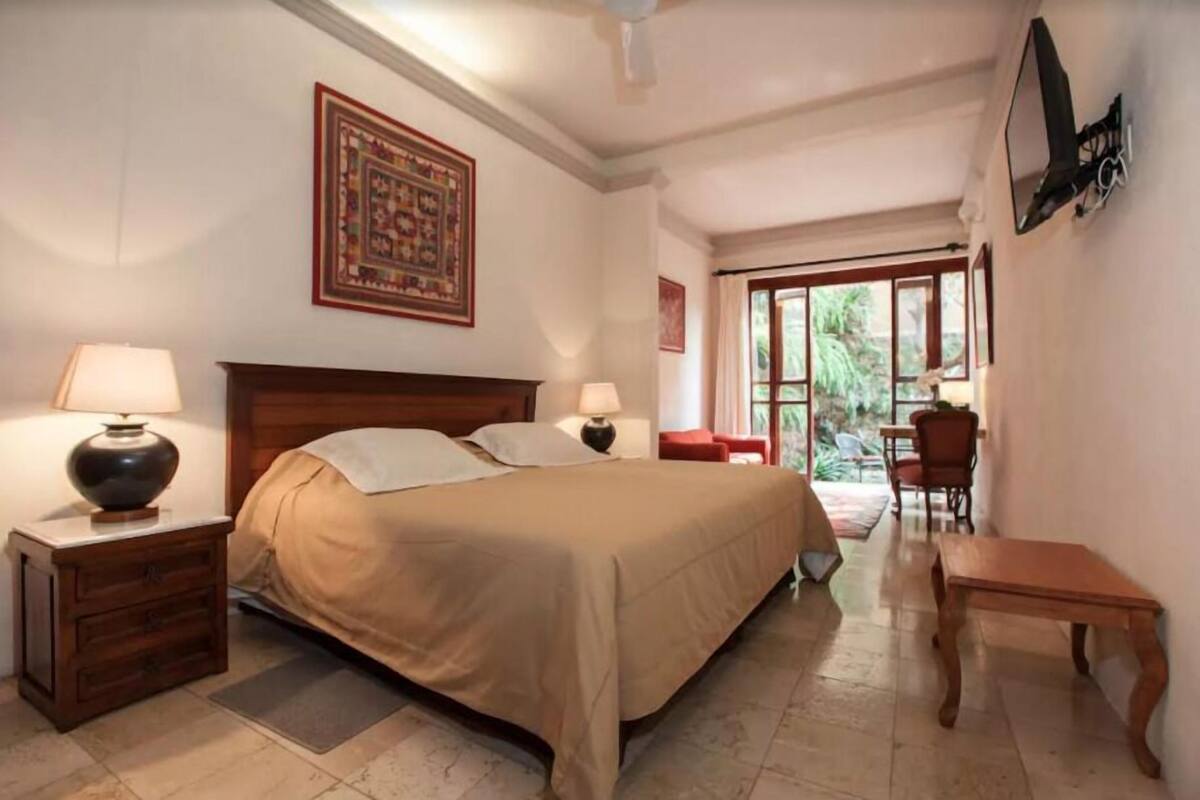 Une chambre à coucher avec un lit, deux tables de chevet avec des lampes, une petite table en bois, une chaise et une vue sur l’extérieur grâce à une porte vitrée.