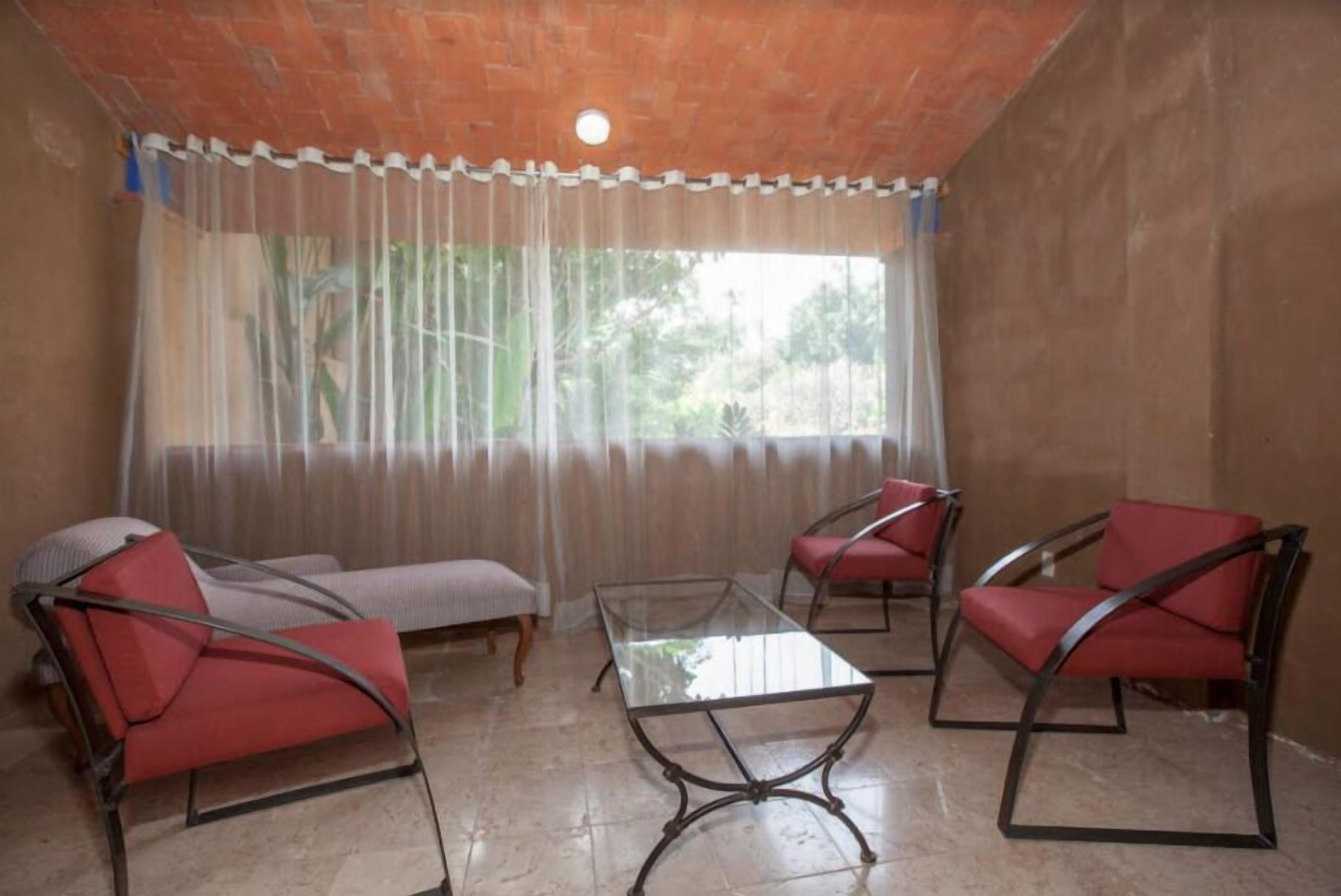 Una habitación con una mesa de vidrio, sillas tapizadas en rojo, un banco y vistas a través de una ventana con cortinas translúcidas.
