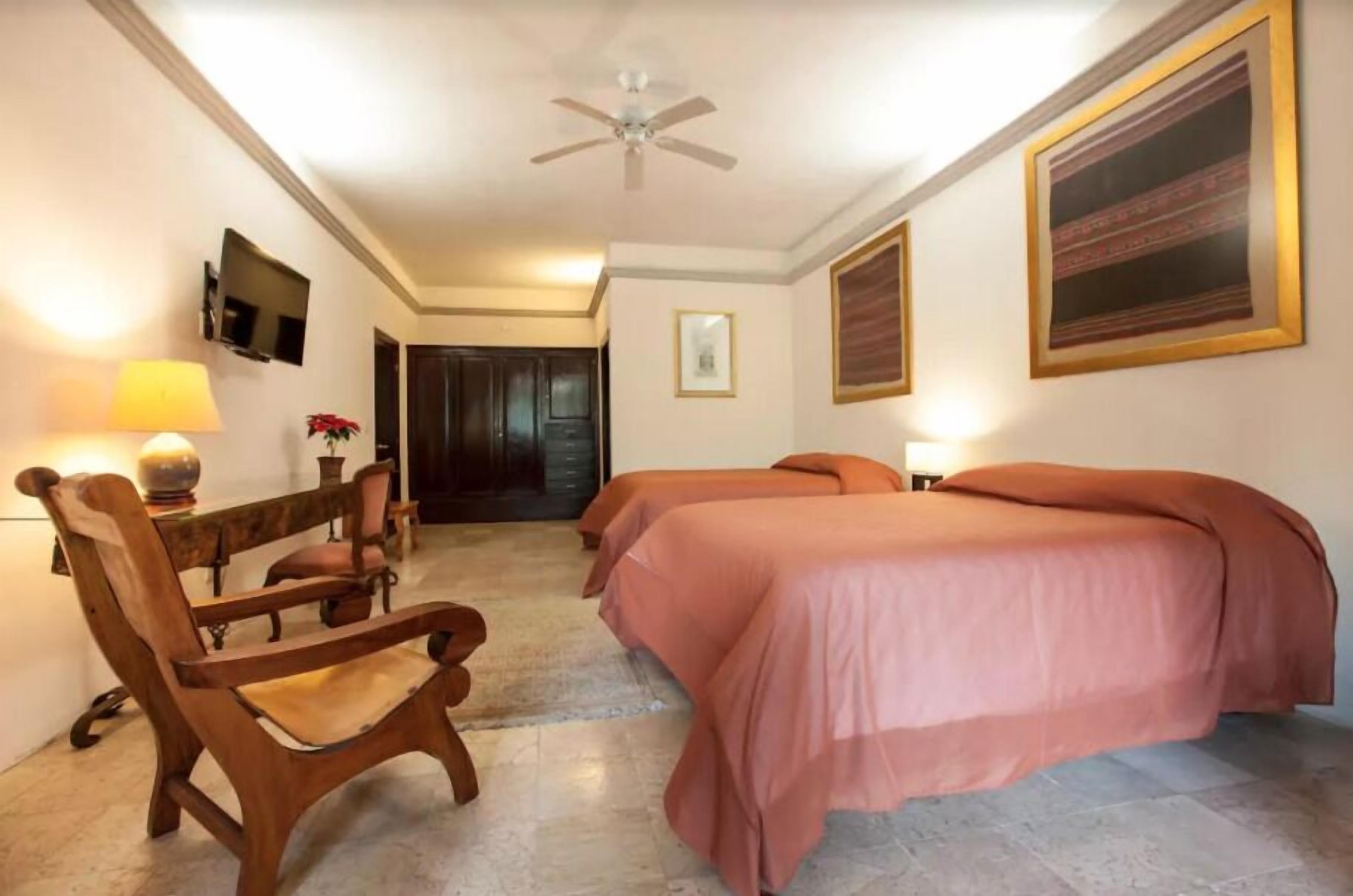 Habitación de hotel con dos camas, una silla de madera, un escritorio con lámpara, televisor y cuadros en las paredes.