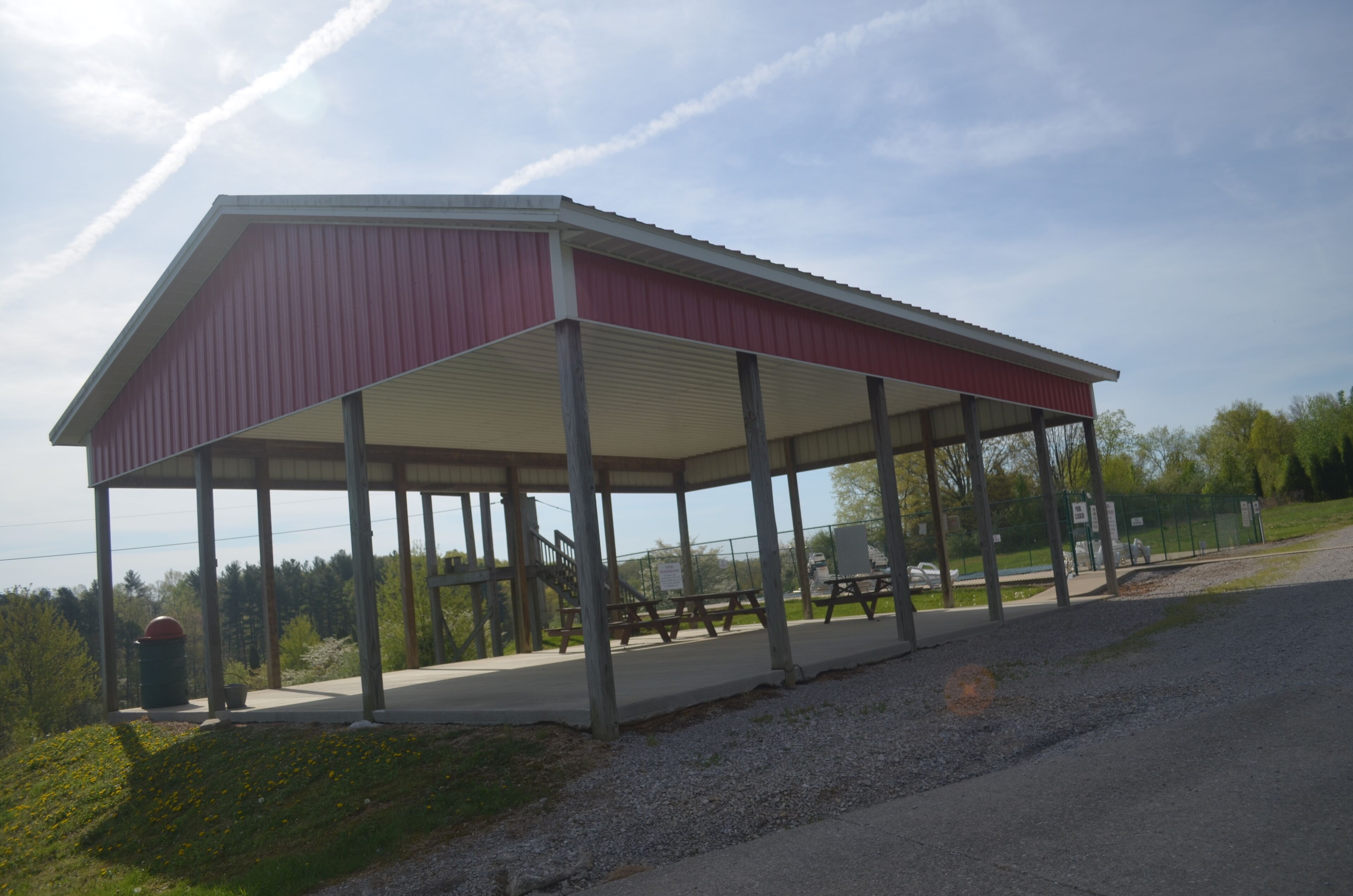 A covered picnic area with benches and picnic tables, surrounded by trees and a gravel path.