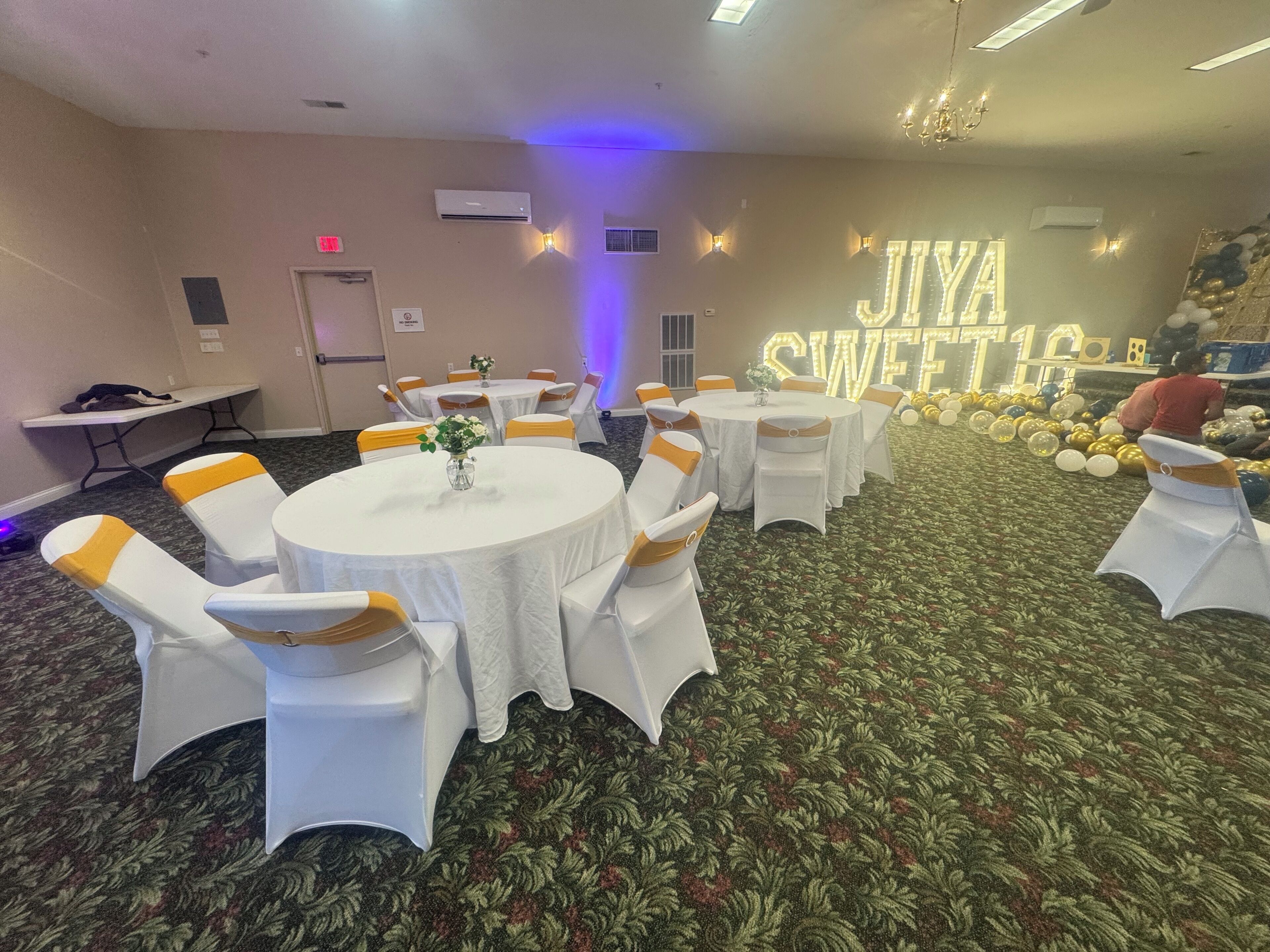 A banquet hall with round tables, white tablecloths, and yellow sashes. There are air conditioning units on the walls, and a large illuminated sign that reads "JIYA SWEET."