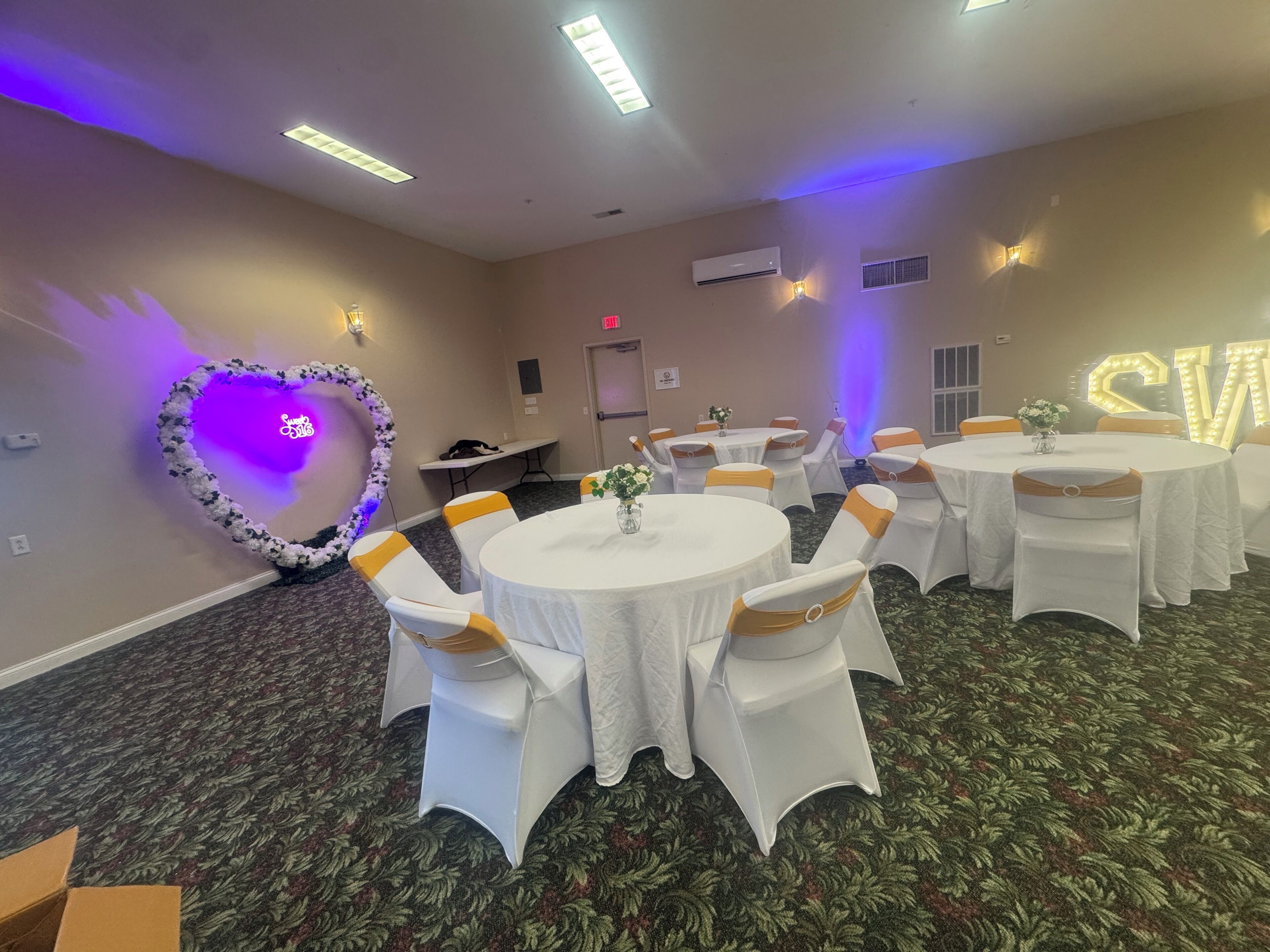 Un salón de conferencias con mesas redondas, sillas blancas con cintas amarillas, un arreglo floral en forma de corazón y un letrero de neón.