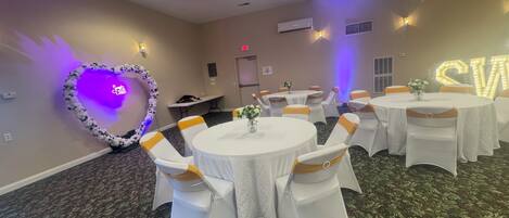 A conference room with round tables, white chairs with yellow sashes, a heart-shaped floral arrangement, and a neon sign.
