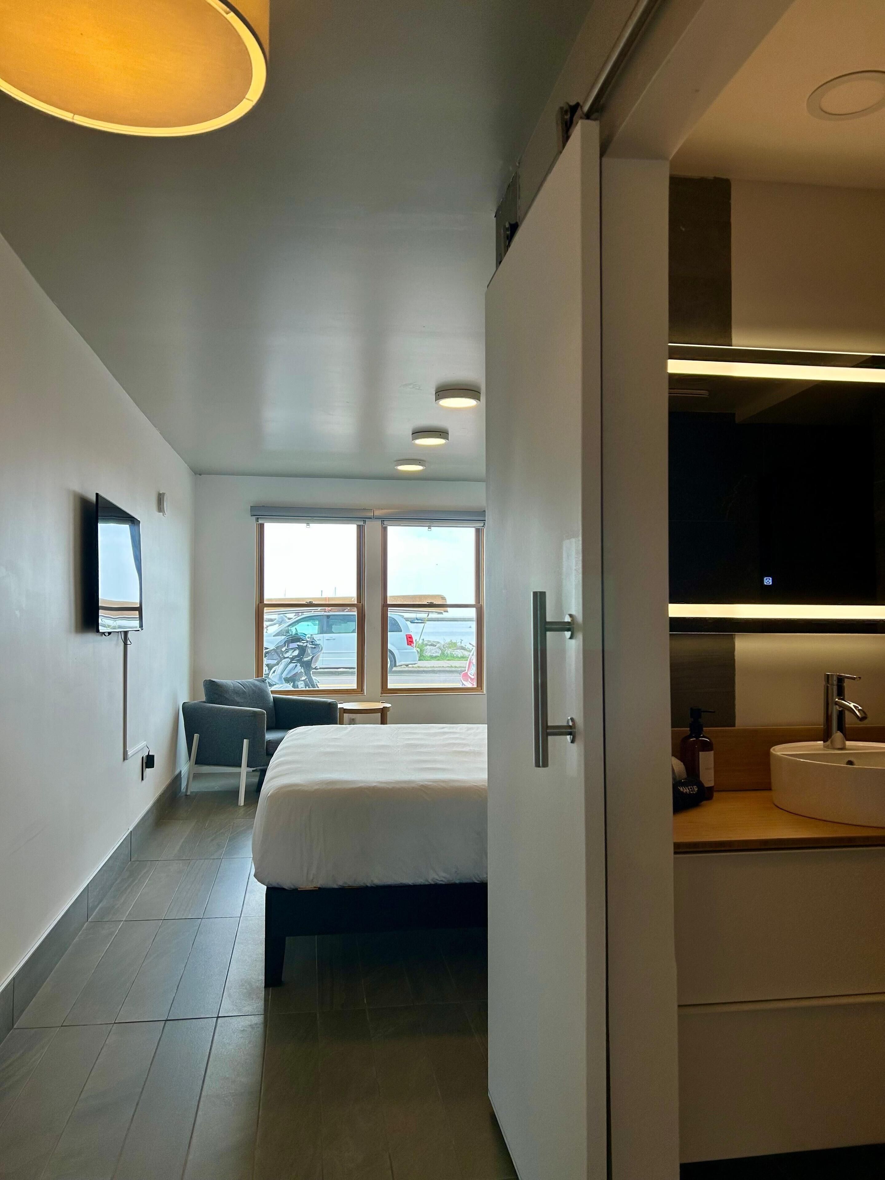 Una moderna camera d'albergo con un letto, una TV a parete, una finestra vista esterna e un bagno con lavandino e specchio.