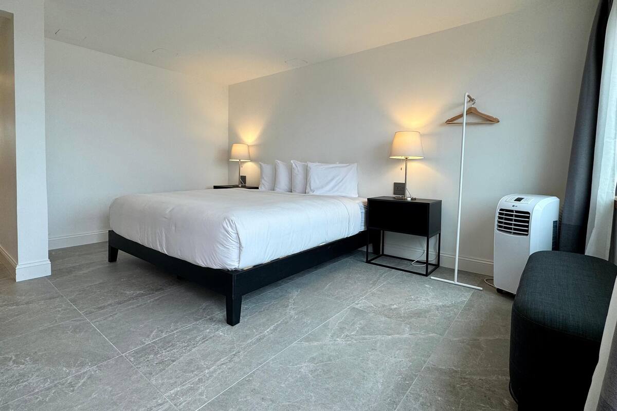 A hotel room with a bed, two bedside tables with lamps, a chair, a heater, and a coat rack.