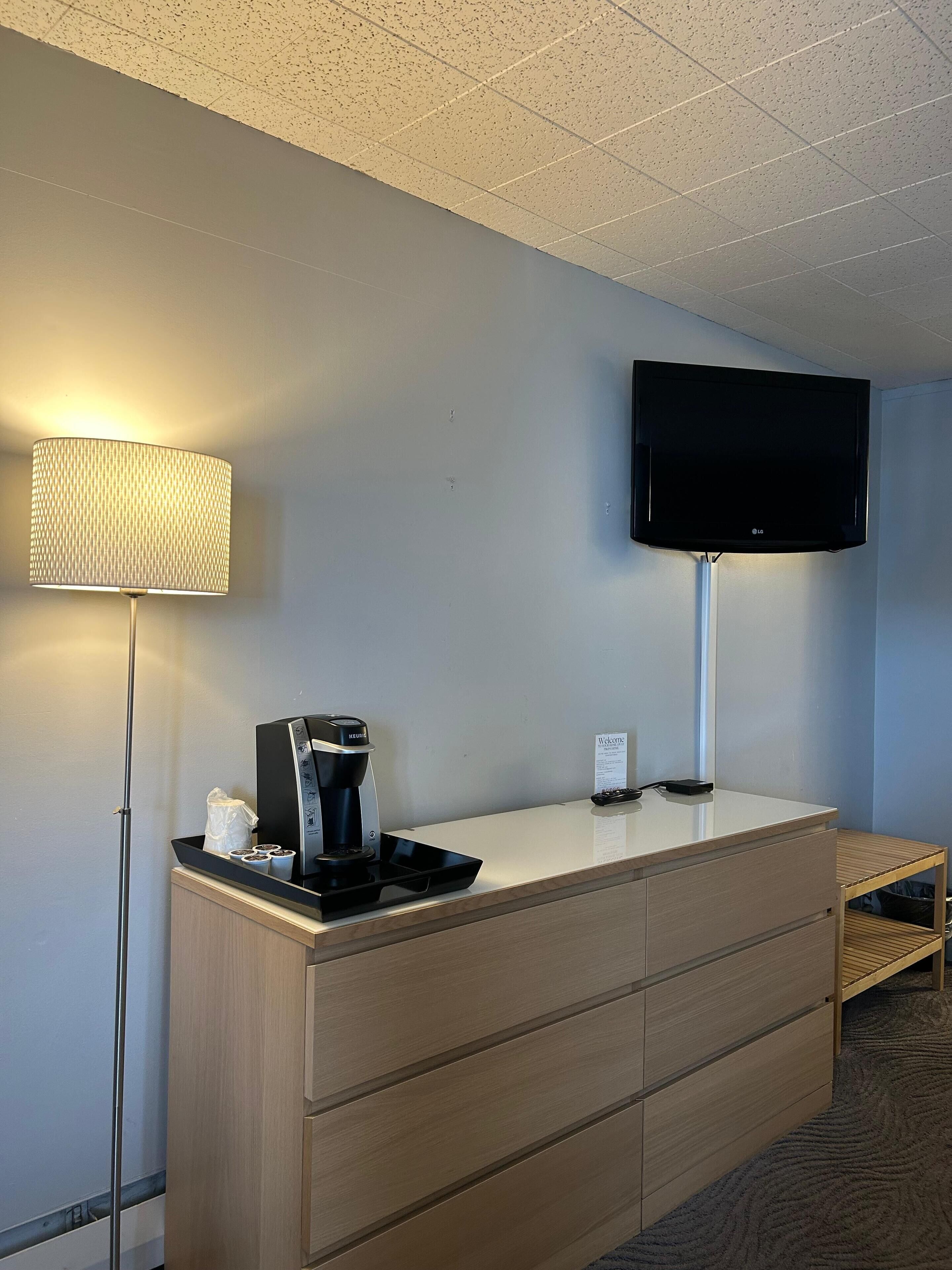 Ett hotellrum med en platt-TV som är monterad på väggen, en kaffebryggare på en bänk och en lampa på ett golvstativ.