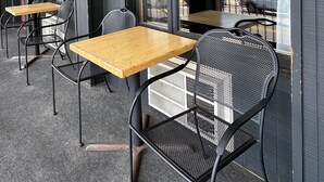 A covered outdoor seating area with tables and chairs, large windows offering a view of the sea and a distant bridge.