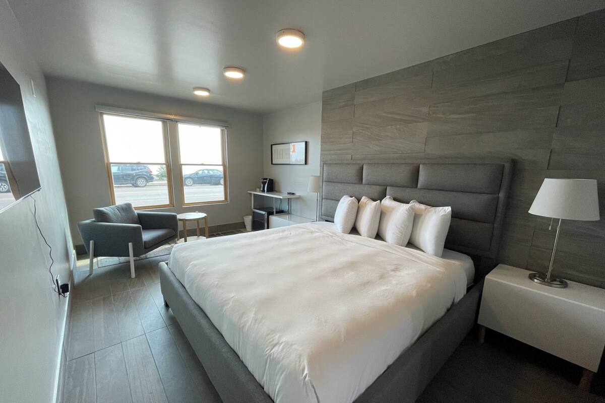 A modern hotel room with a large bed, a desk, a chair, and a view of the street through the window.