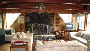 A cozy living room with a stone fireplace, wooden ceiling, and large windows. - Kern Riverfront Lodge (Kernville)