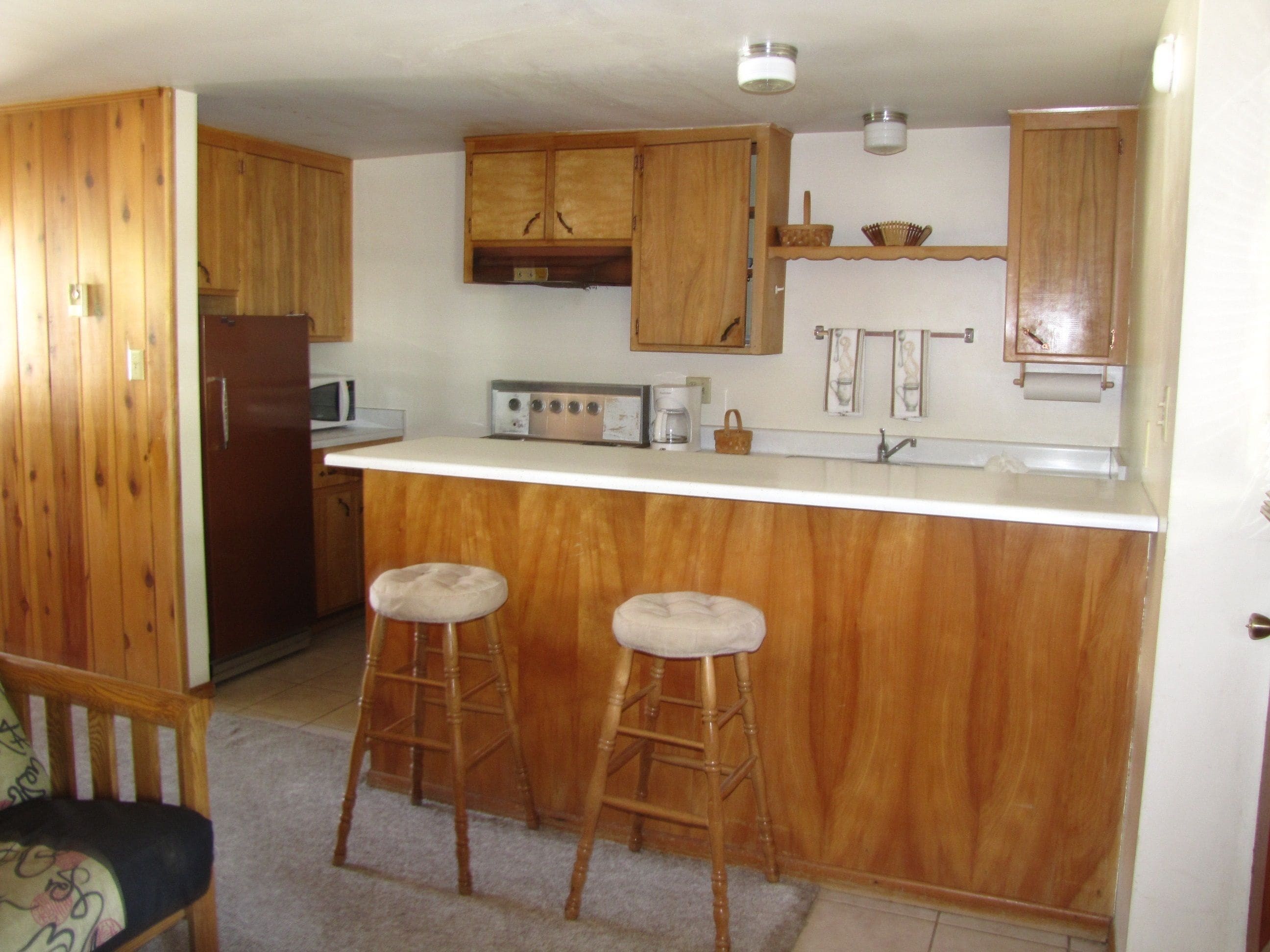 A compact kitchen with wooden cabinets, a microwave, a stove, and two stools.