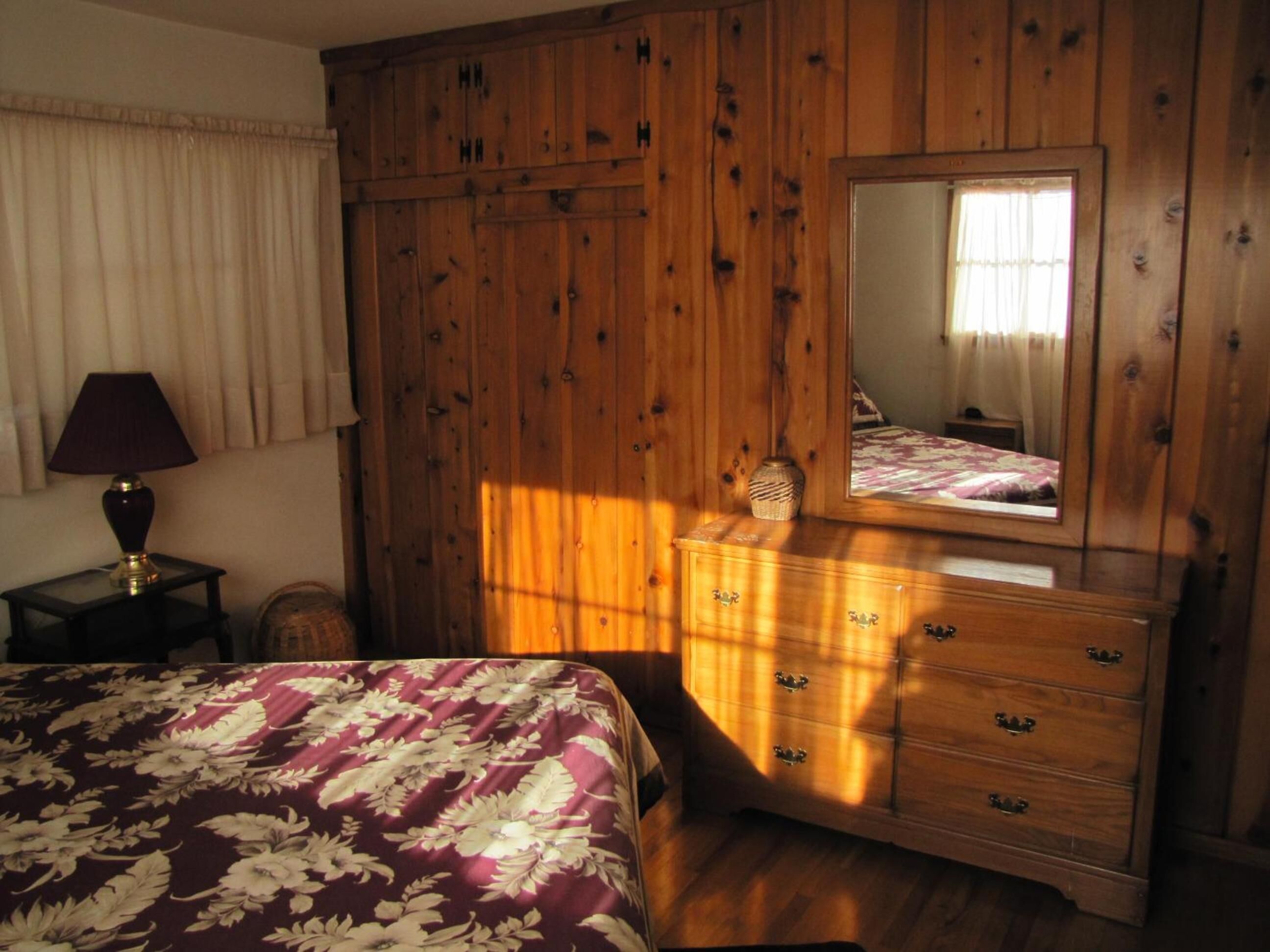A bedroom with a wooden wardrobe, a bed with a floral bedspread, a bedside table with a lamp, and a mirror above the dresser.