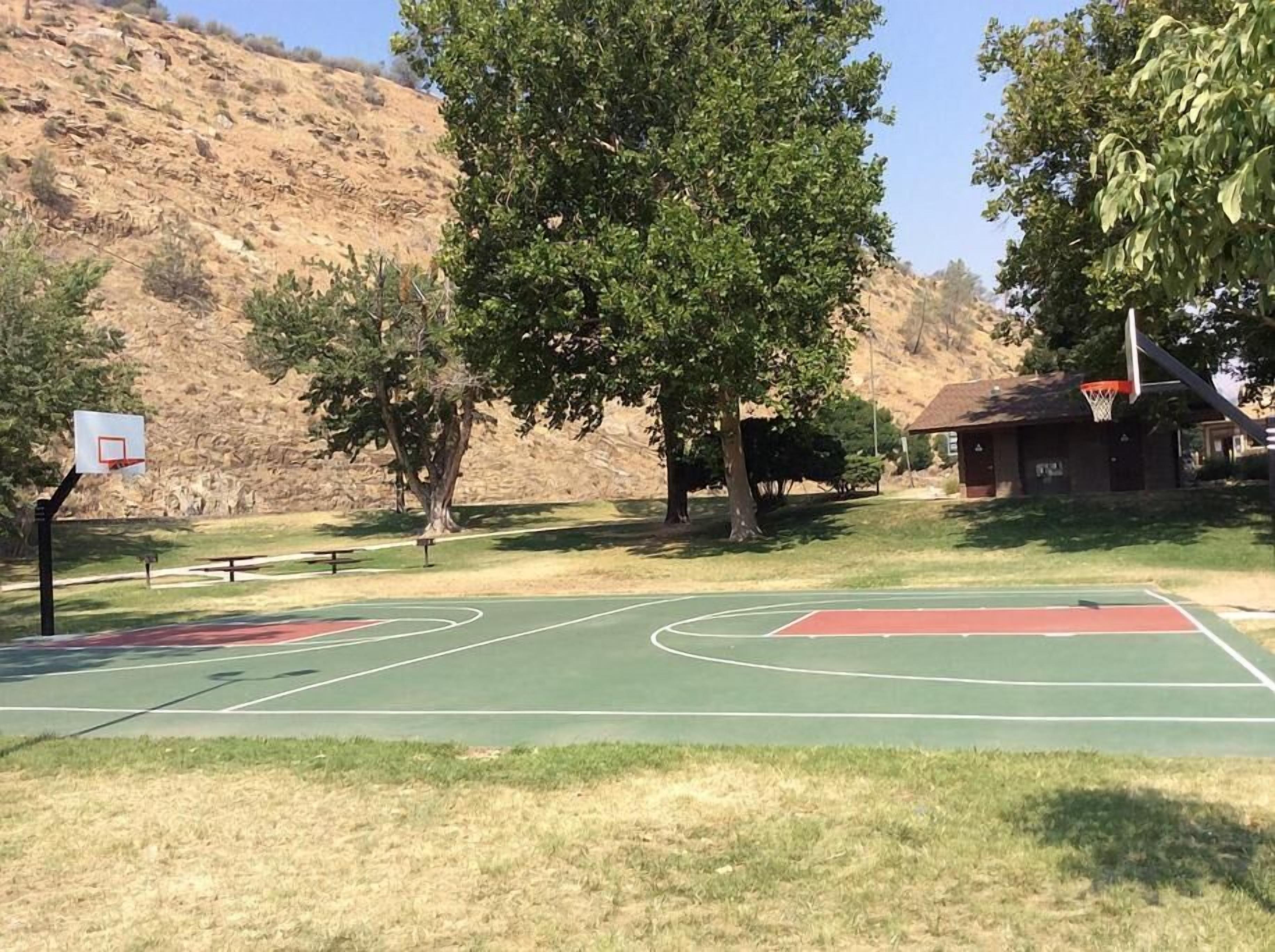 An outdoor basketball court with two hoops, surrounded by trees and a hillside in the background.