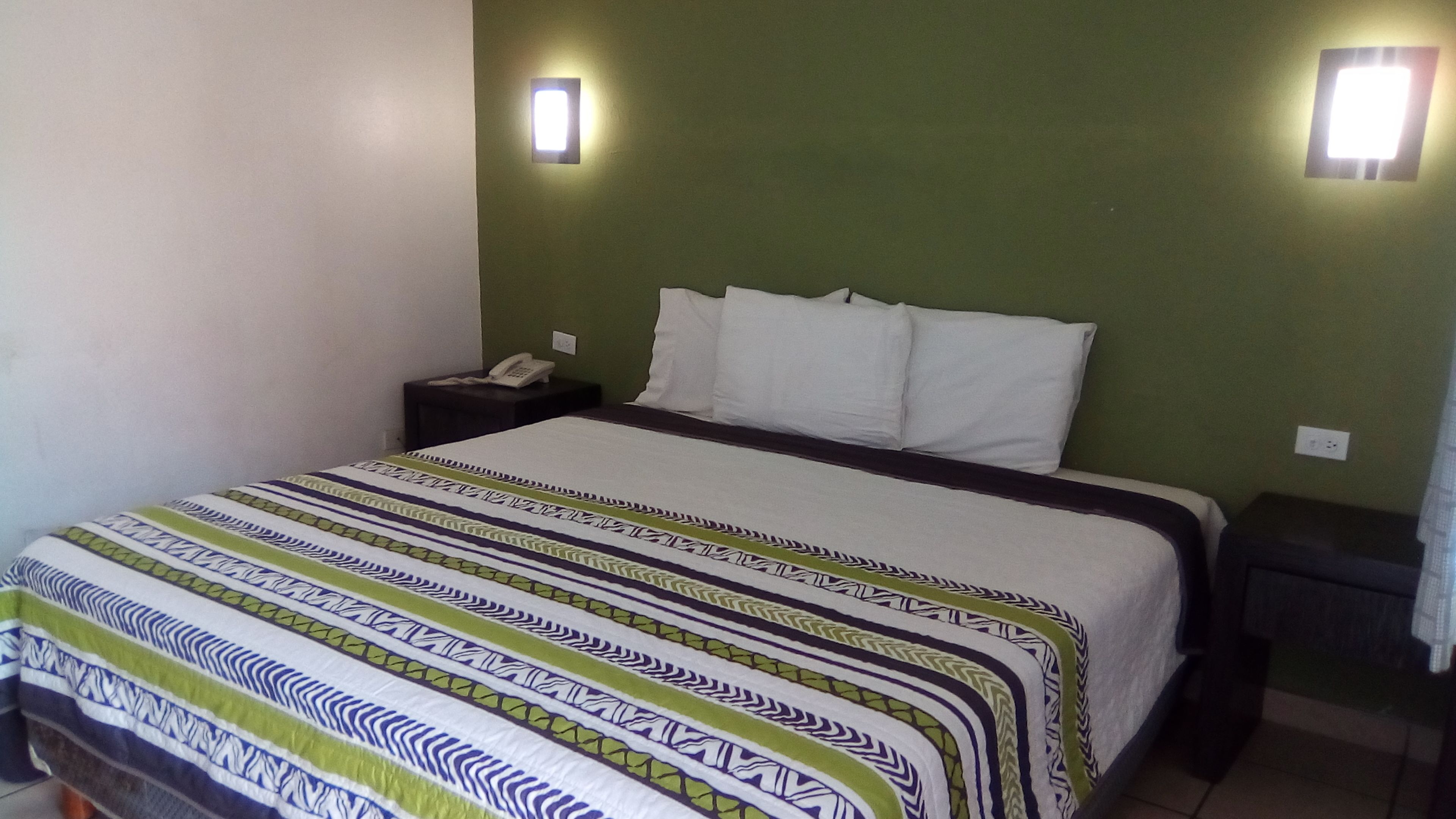 Una habitación de hotel con una cama, una mesita de noche, un teléfono y dos lámparas fijadas a la pared.