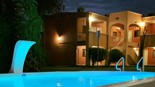 Una piscina amb un element d'il·luminació modern, un edifici amb múltiples balcons i il·luminació càlida, i una zona de jardí ben cuidada.