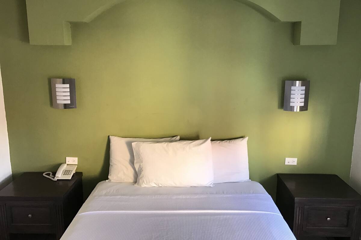 Una habitación con una cama, dos mesitas de noche, un teléfono sobre una de ellas y dos lámparas fijadas en la pared.