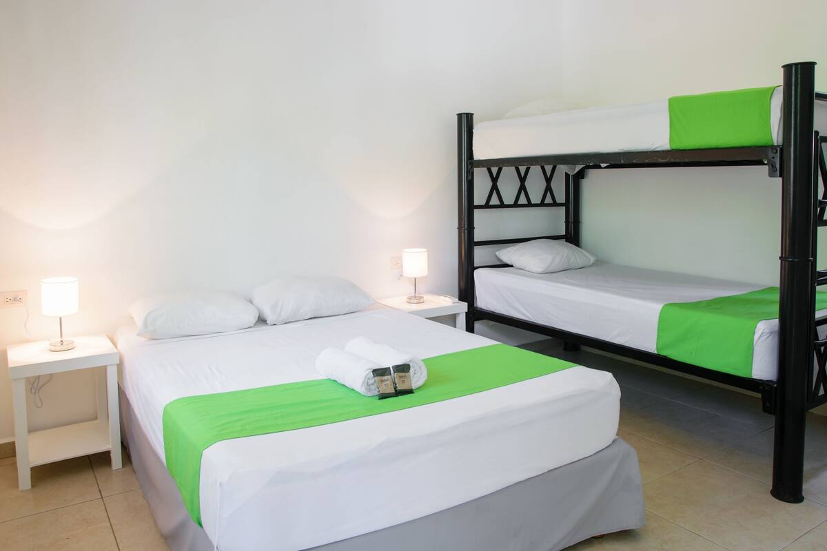 Ein Hotelzimmer mit zwei Betten, davon ein Etagenbett und ein Einzelbett. Zwei Nachttische mit Lampen stehen bereit, und ein grünes Handtuch liegt auf dem Einzelbett.