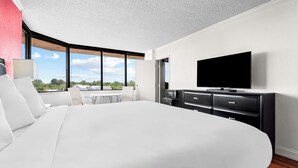 A modern hotel room with a large bed, a flat-screen TV, a dining area with a table and chairs, and a view of the outdoors.