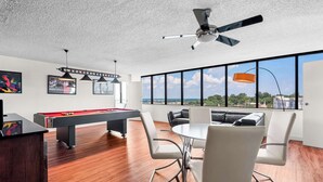 A modern room with a pool table, white chairs, a ceiling fan, and large windows offering a view of the city.