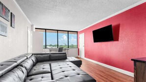 A modern living room with a sectional sofa, a flat-screen TV mounted on a red wall, and a large window offering a view of the outdoors.