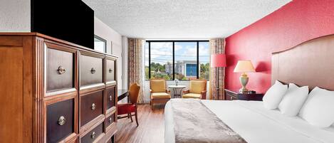 A hotel room with a large bed, a wooden dresser, two chairs, a small table, and a view of the outdoors.