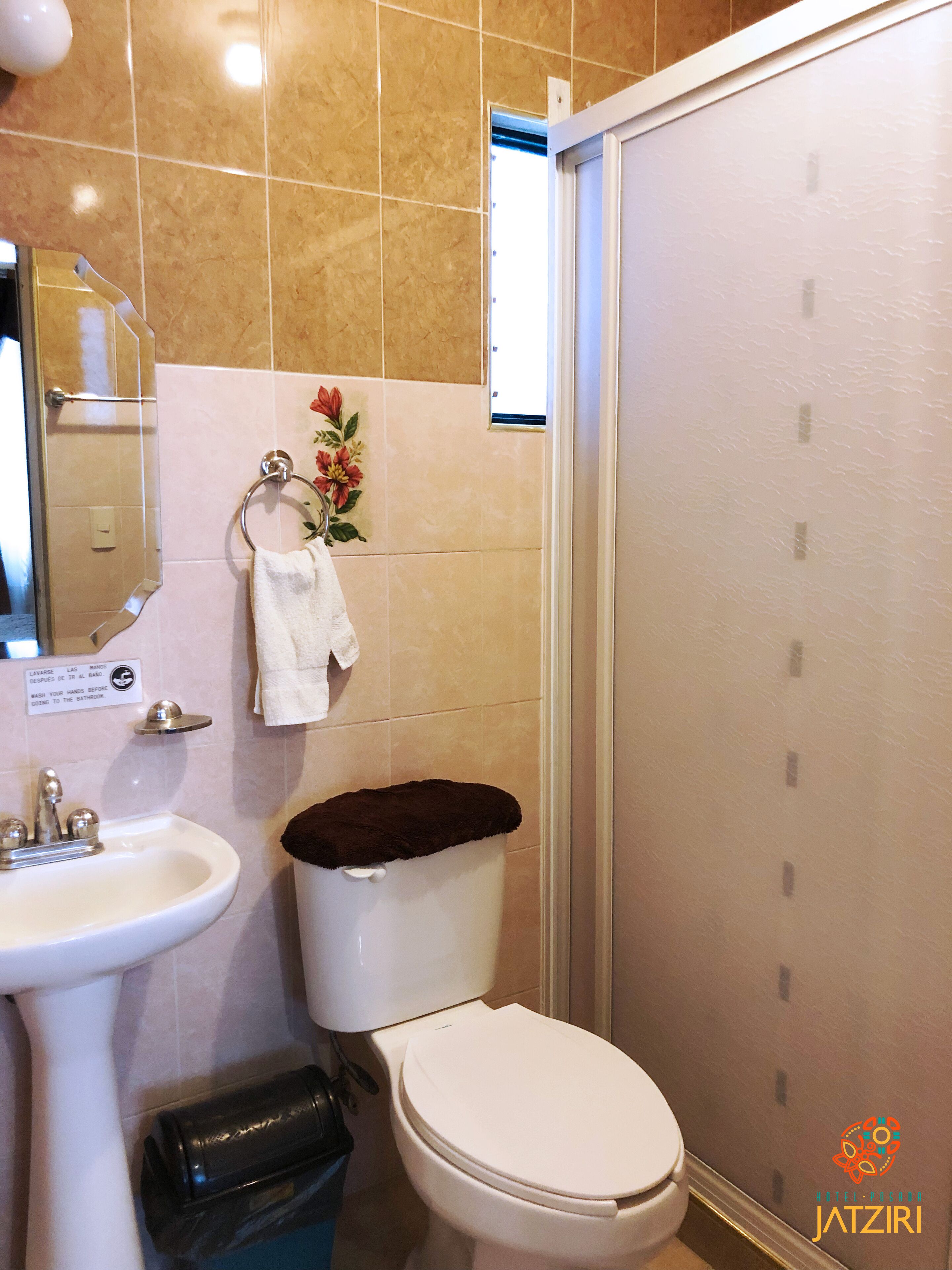 A bathroom with a toilet, sink, and shower. There is a towel hanging on the wall and a trash bin next to the toilet.