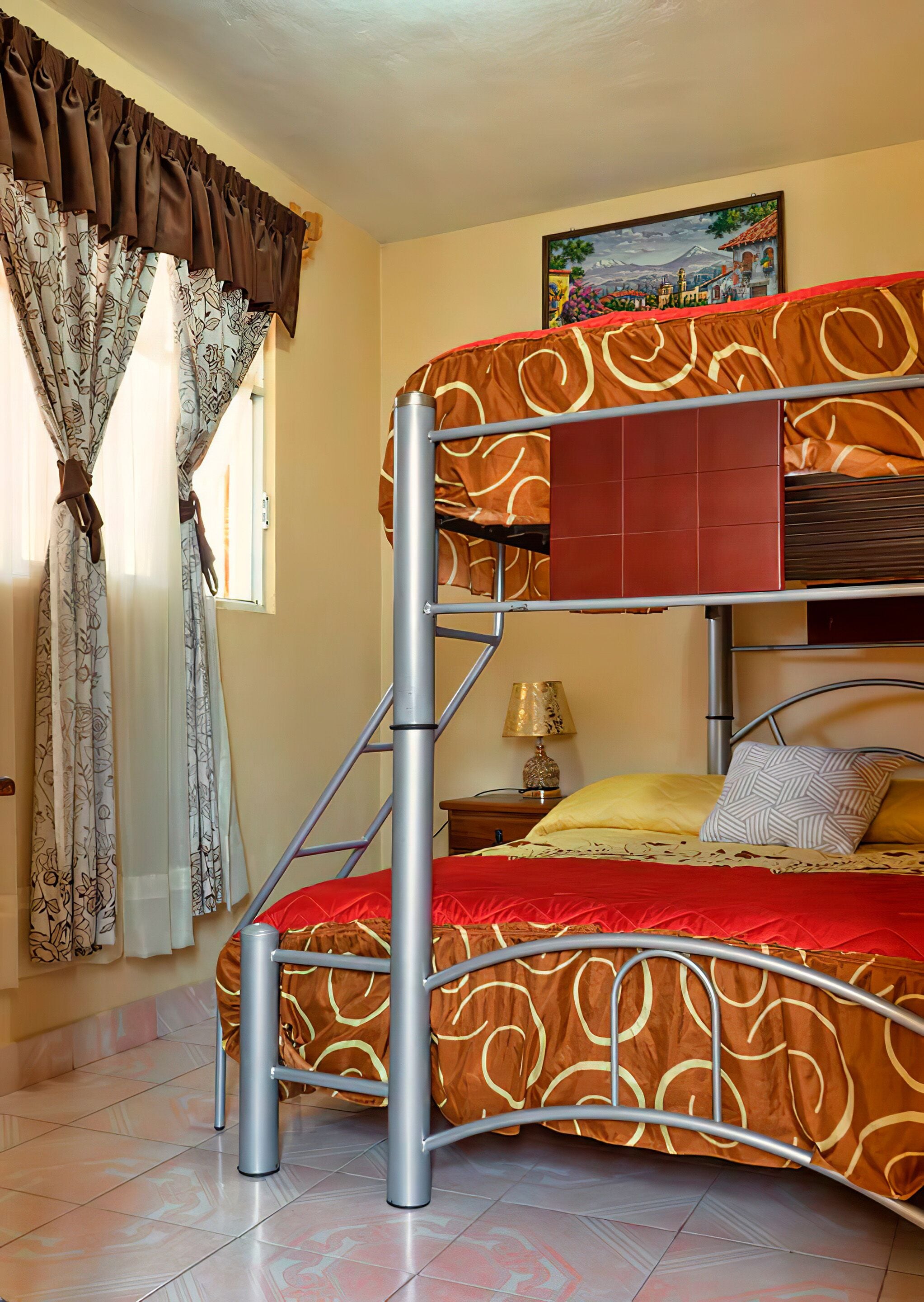 A bunk bed with a metal frame, a red and brown patterned bedspread, a small bedside table with a lamp, and a window with curtains.