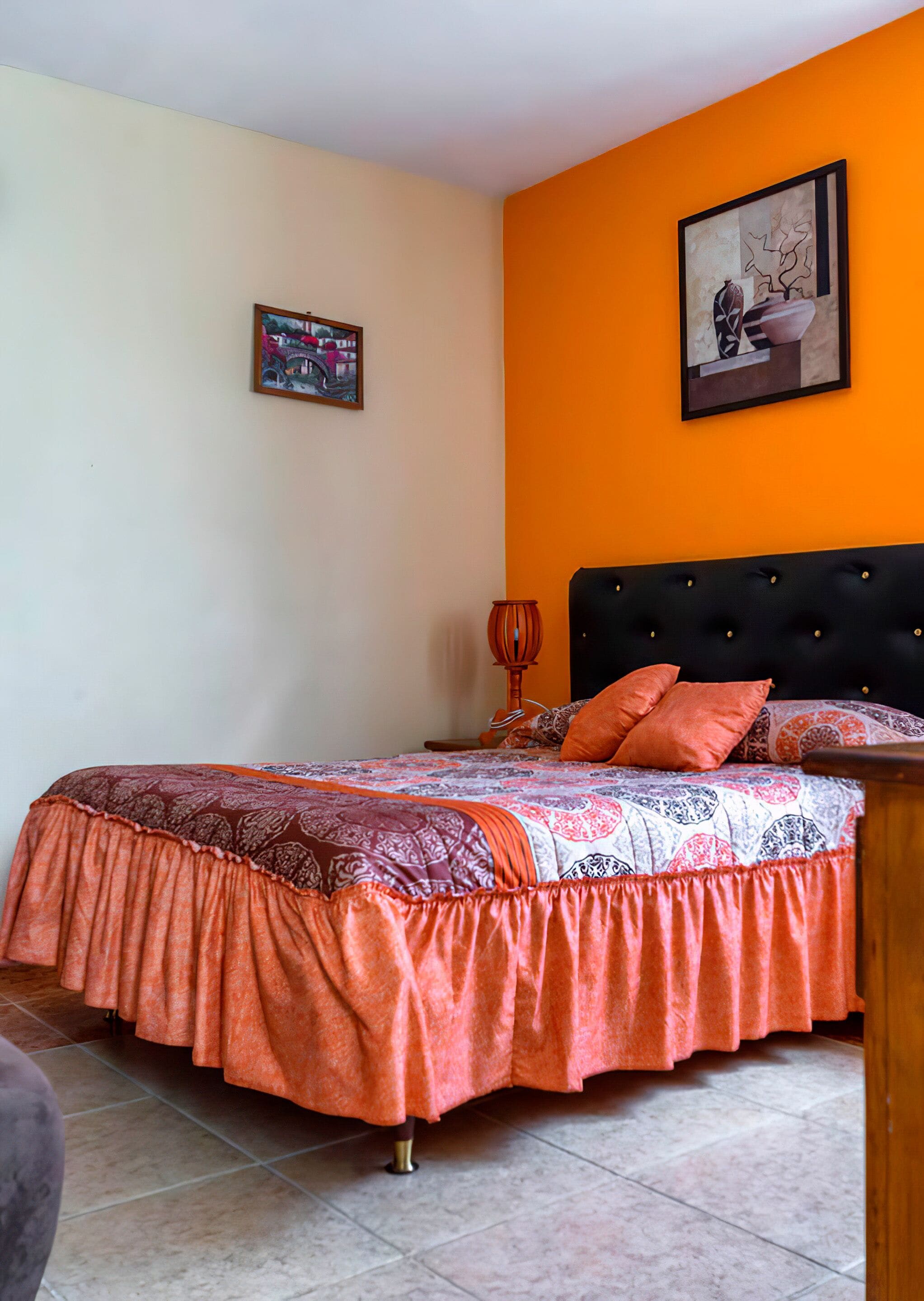 Un dormitorio con cama, cabecera, mesita de noche, lámpara y dos cuadros en la pared.