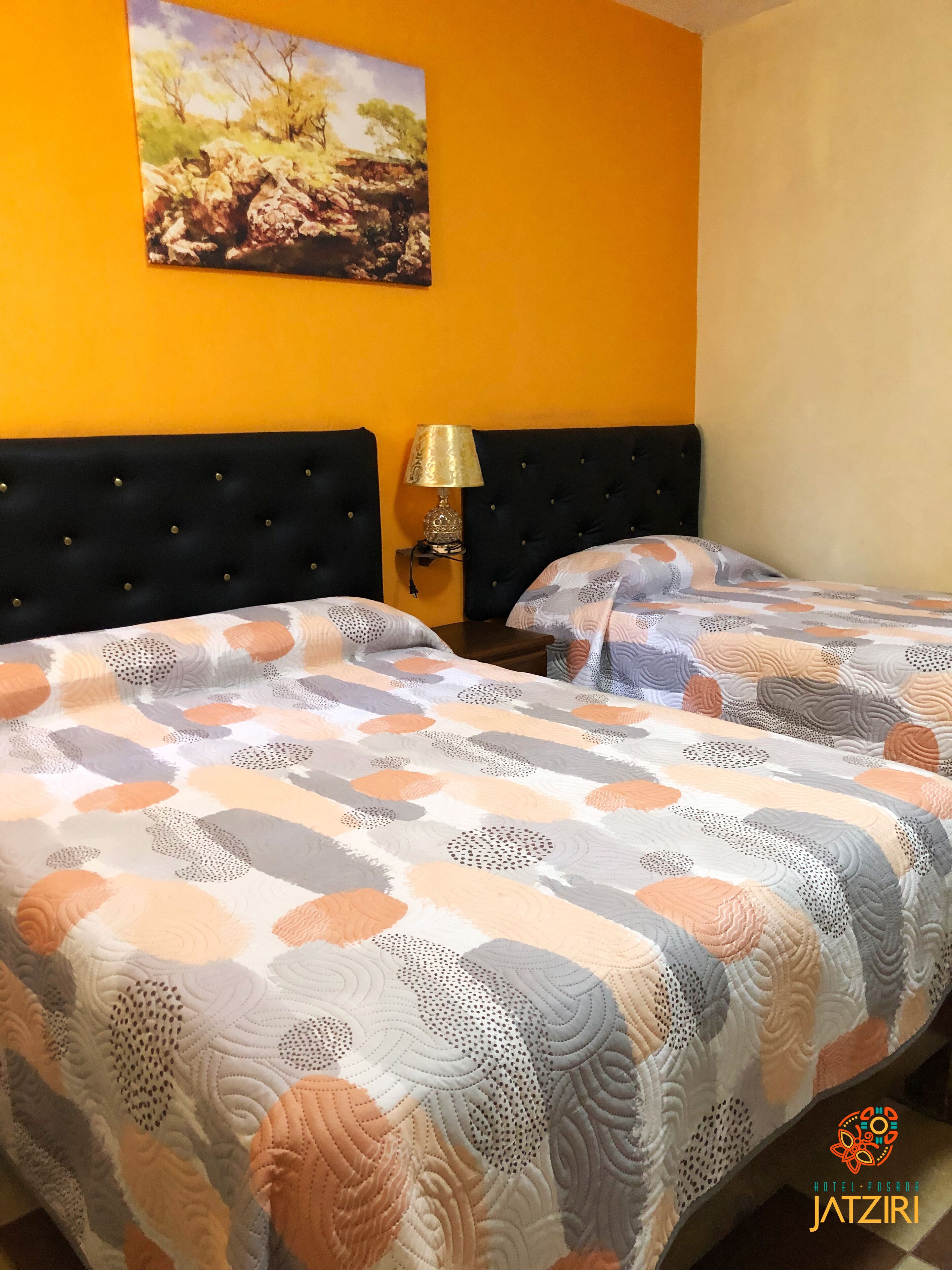 Een tweepersoonsbed met een bedsprei met een patroon, een zwart bekleden hoofdeinde, een nachtkastje met een lamp en een schilderij aan de muur.