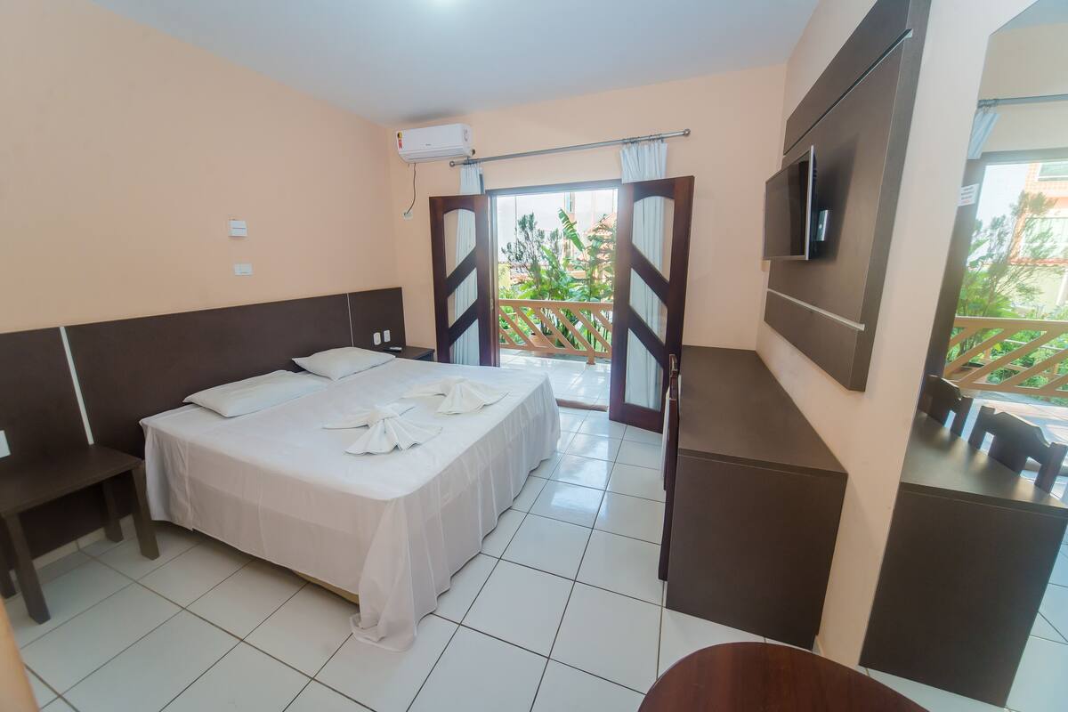 Habitación de hotel con cama, escritorio, televisor y balcón con vistas a zonas verdes.