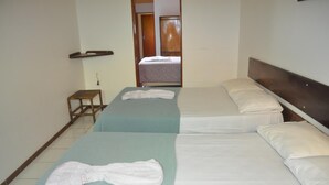 Quarto de hotel com duas camas, cada uma com colcha azul e roupa de cama branca. Há um banquinho de madeira pequeno e uma estante acima das camas.