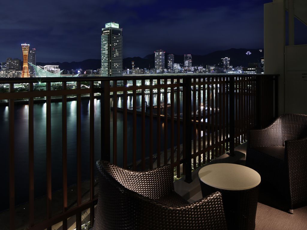 Un balcon agrémenté de meubles en osier, une vue sur les lumières d’une ville la nuit, et un plan d’eau.