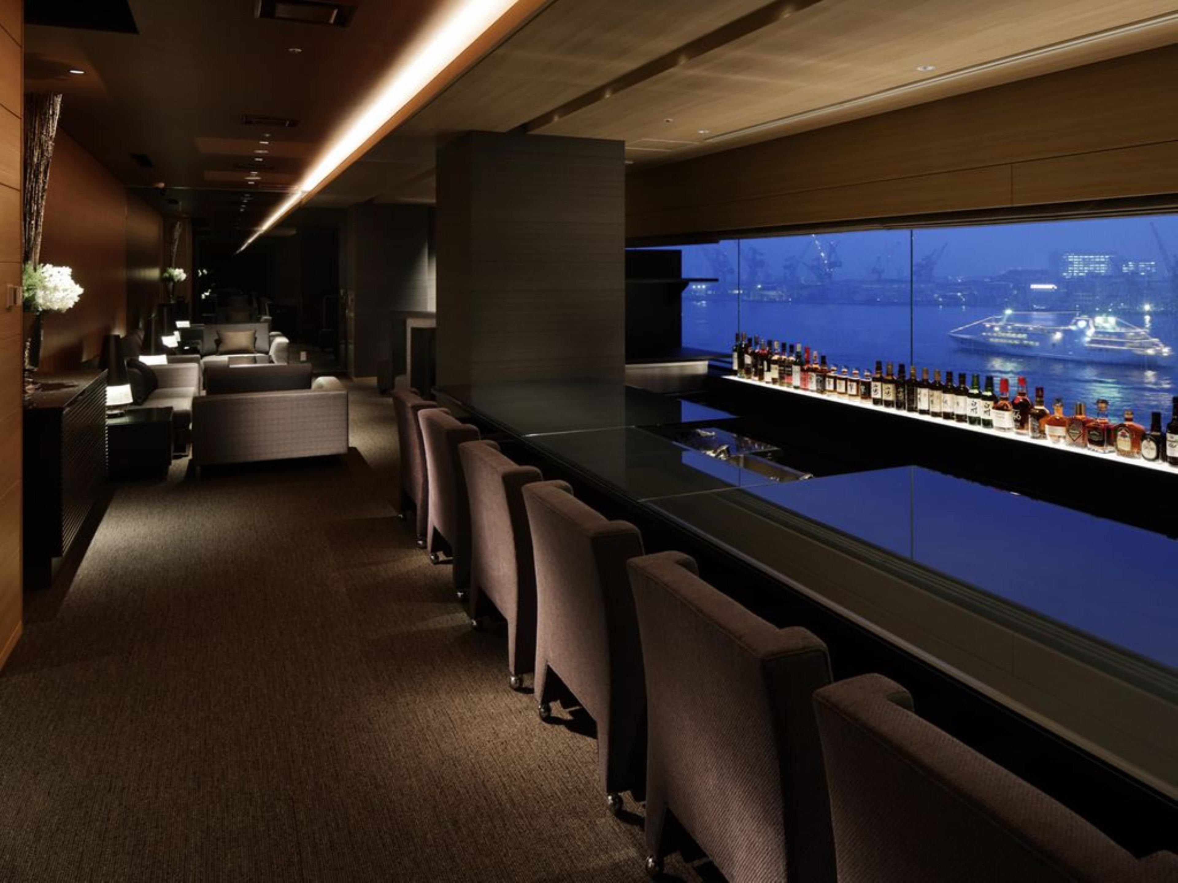 A bar area with a long counter, multiple seating options, and a large window with a view of a body of water and ships.