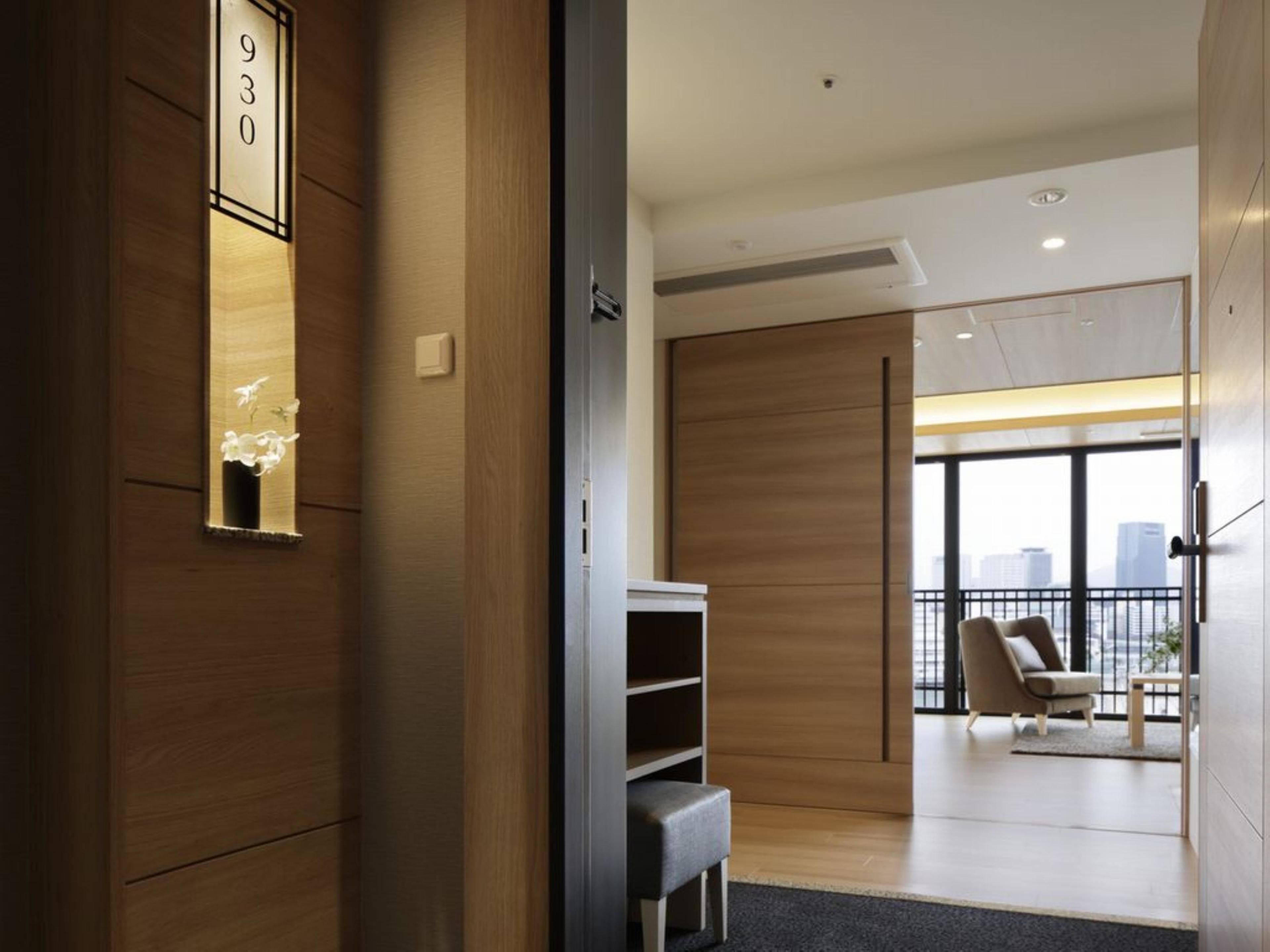 A modern hotel room with a wooden interior, a large window with a city view, a seating area with a chair, and a small table.