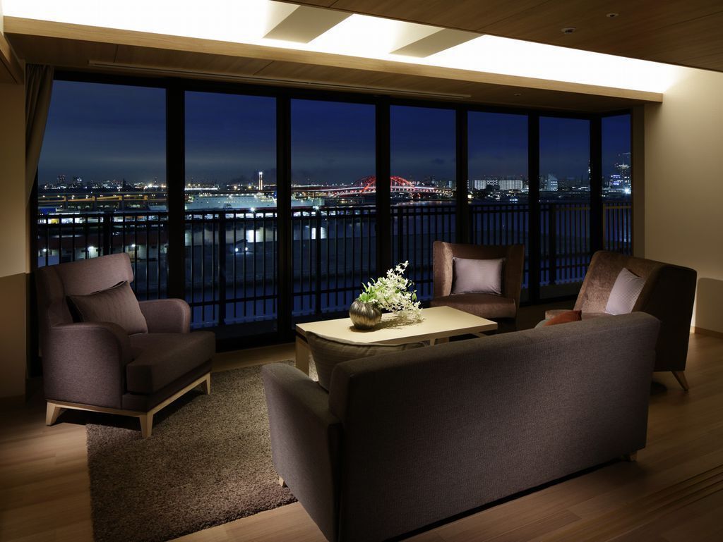 Un salon moderne doté d’une grande fenêtre donnant sur une vue panoramique de la ville la nuit, avec un canapé, des fauteuils et une table basse.