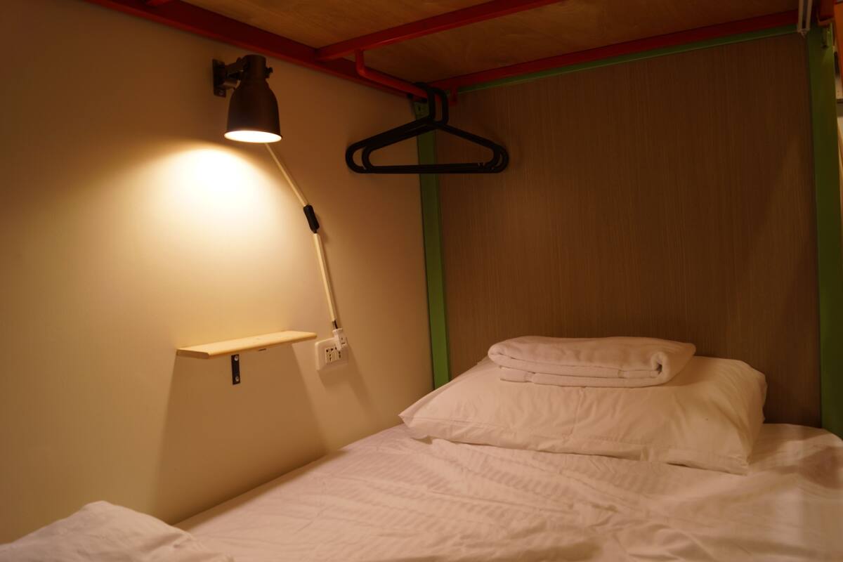 A single bed with white bedding, a wall-mounted lamp, and a hanging garment rack.