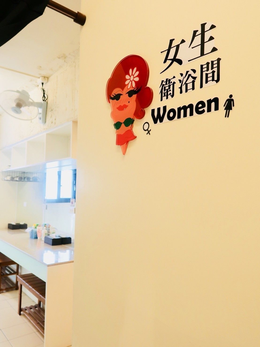 Eine Wand mit einem dekorativen Schild, das eine stilisierte Frauenfigur zeigt und Text in Chinesisch und Englisch enthält, der ein Damentoilette-Hinweis darstellt.