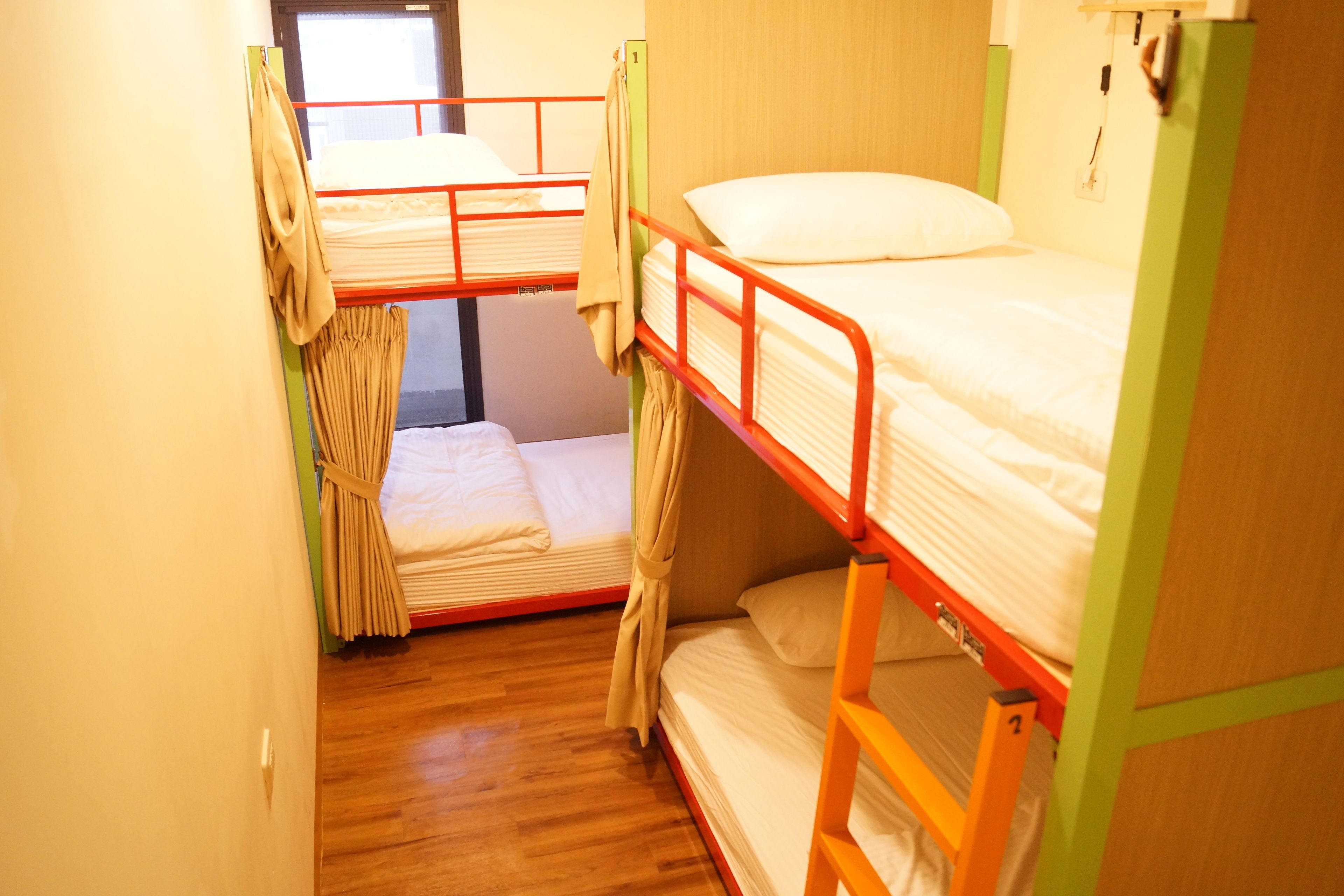 A dormitory room with bunk beds, wooden flooring, and a window with curtains.