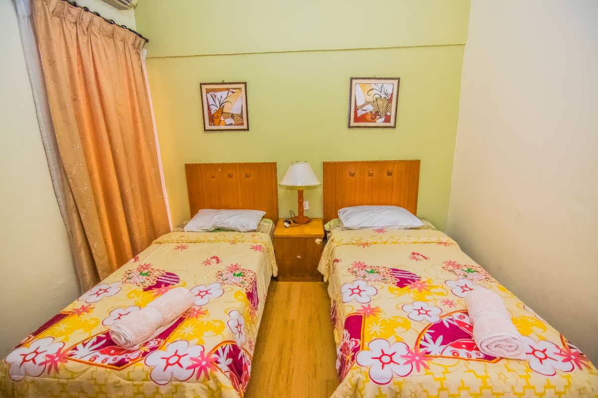 A room with two single beds, each with a floral bedspread and a rolled towel. There is a nightstand with a lamp between the beds.