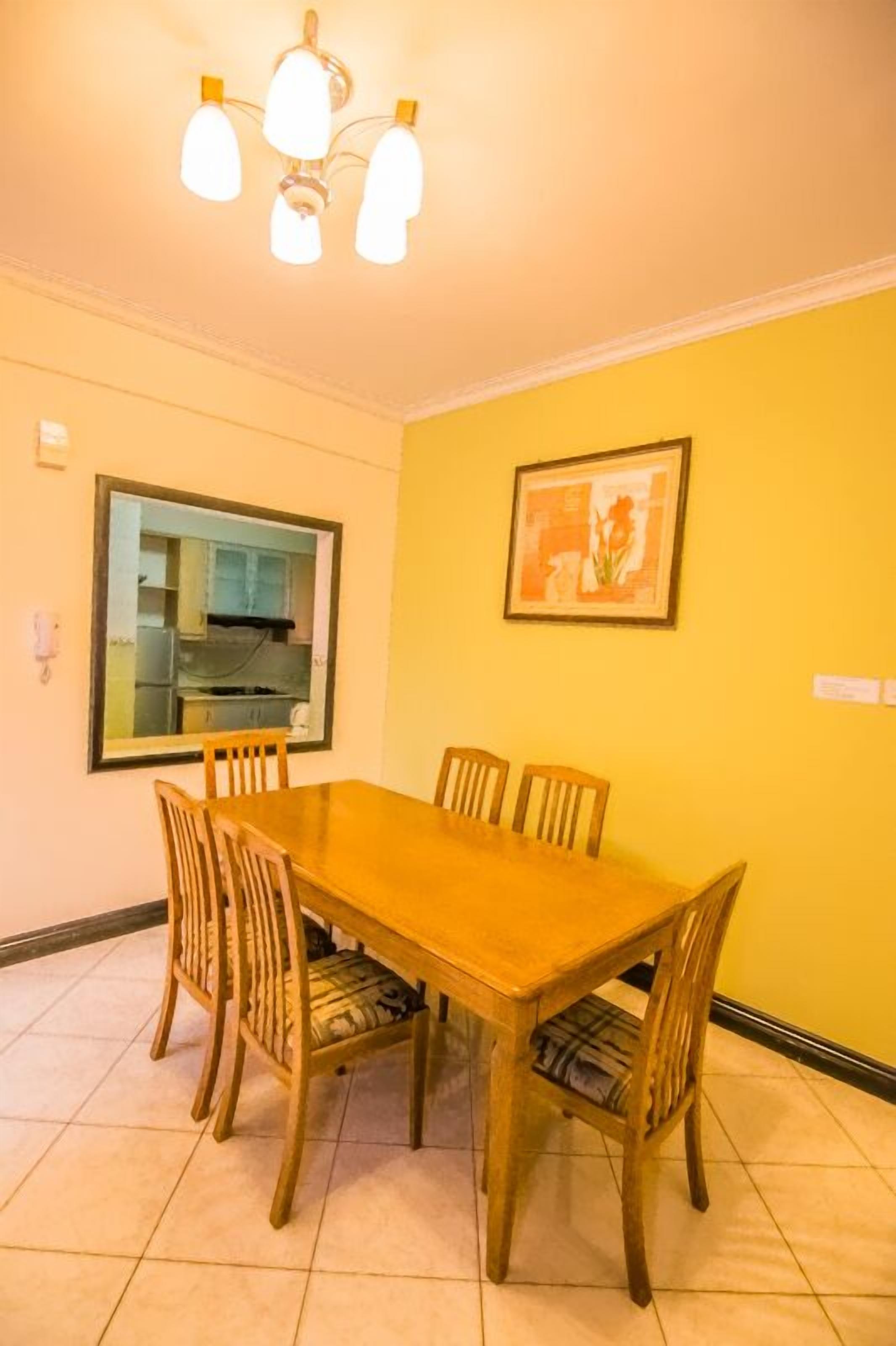 A dining area with a wooden table and chairs, a kitchen visible through a window, and a ceiling light fixture.