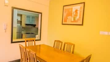 A dining area with a wooden table and chairs, a kitchen visible through a window, and a ceiling light fixture.